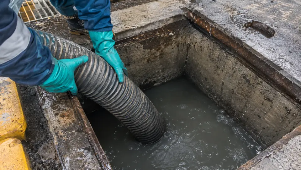 Manos con guantes bajando una manguera de succión a una cámara de hormigón con agua turbia